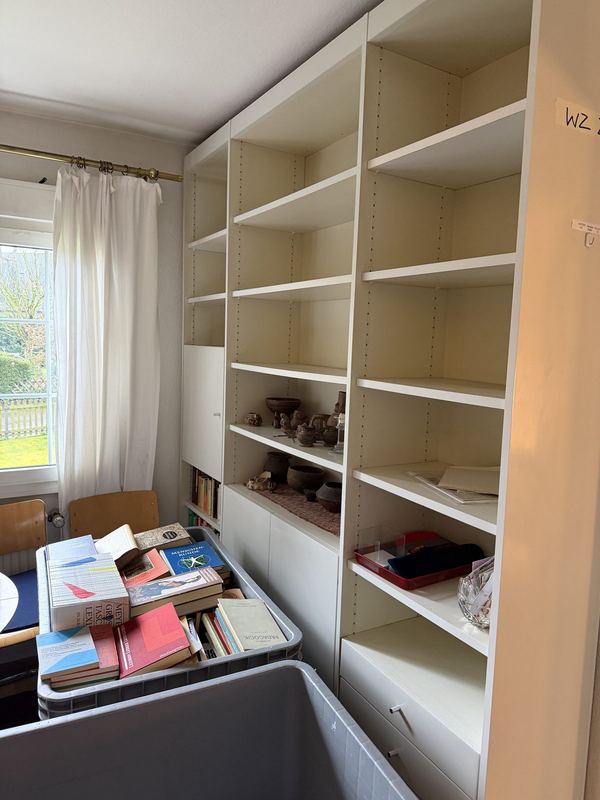 Nahezu leeres Bücherregal Weißes Bücherregal, nahezu vollständig geleert, nur einzelne Gegenstände stehen noch darin, davor ein grauer Rollbehälter voller Bücher