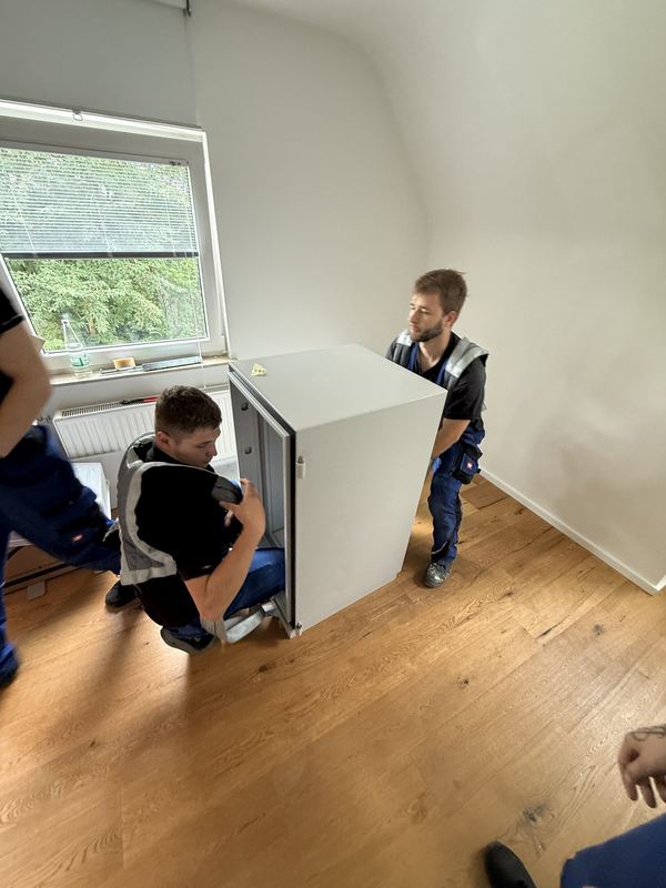 Ein weißes Einbaugerät wird von zwei Mitarbeitern durch ein leergeräumtes Zimmer getragen