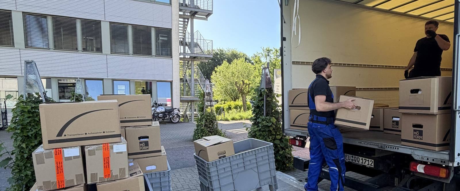 So arbeiten wir bei Umzügen in Meerbusch: Vor einem Bürogebäude mit Außentreppe steht ein Umzugswagen mit offenem Laderaum, ein Mitarbeiter trägt einen Karton zur Ladebordwand, ein zweiter steht im Laderaum, im Vordergrund gestapelte Umzugskartons und graue Kunststoffboxen.