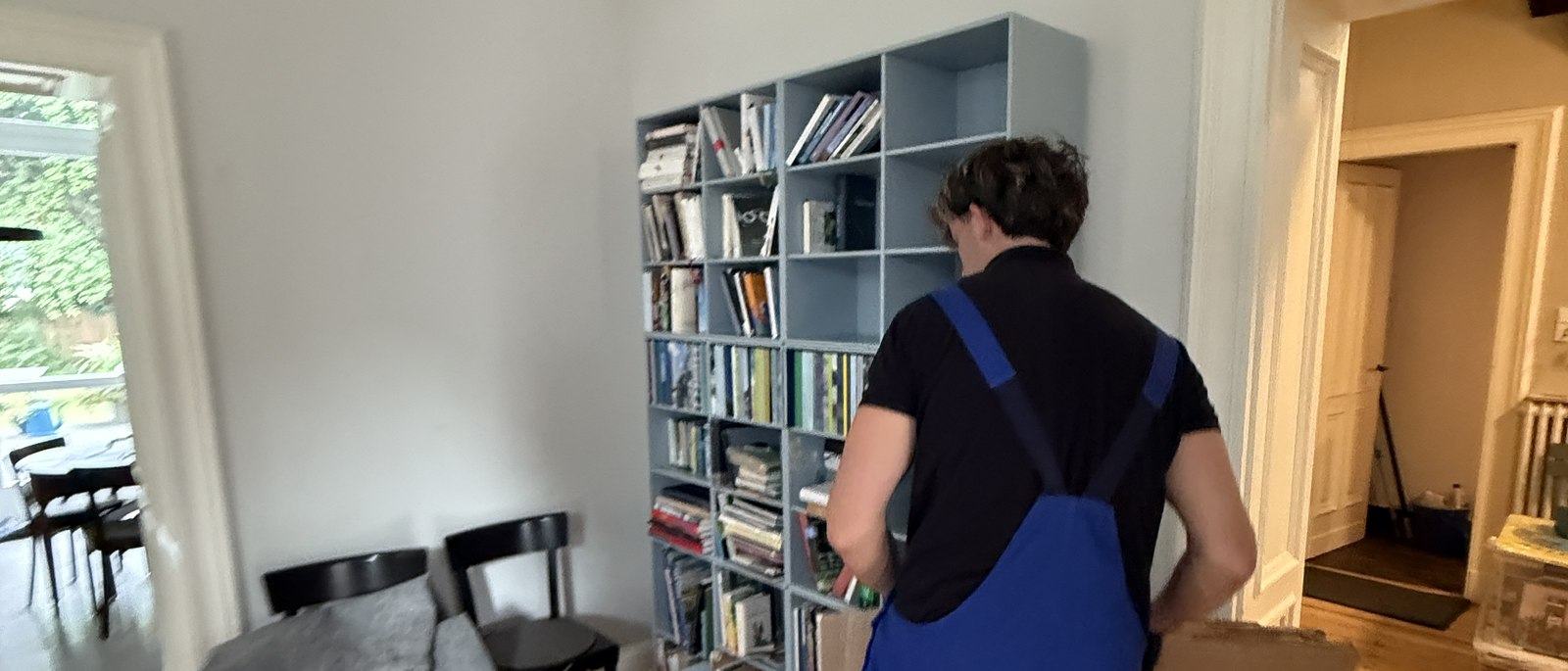 Ihre Fragen zum Umzug in Meerbusch, unsere Antworten: Ein Mitarbeiter in Latzhose steht vor einem hellgrauen Würfelregal mit Büchern und Ordnern und packt dessen Inhalt in einen Karton, im Vordergrund ein Esstisch mit Stühlen.