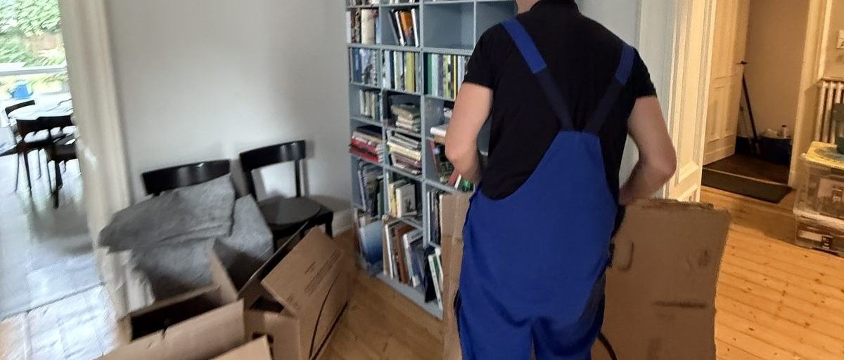Haushaltsauflösung in Meerbusch: Ein Mitarbeiter mit Latzhose trägt einen flach gefalteten Karton vor einem vollen Bücherregal entlang, davor Stühle und weitere Kartons