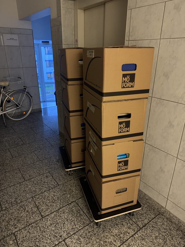 Ein Rollbrett mit vier übereinander gestapelten Kartons steht im gefliesten Eingangsflur eines Hauses