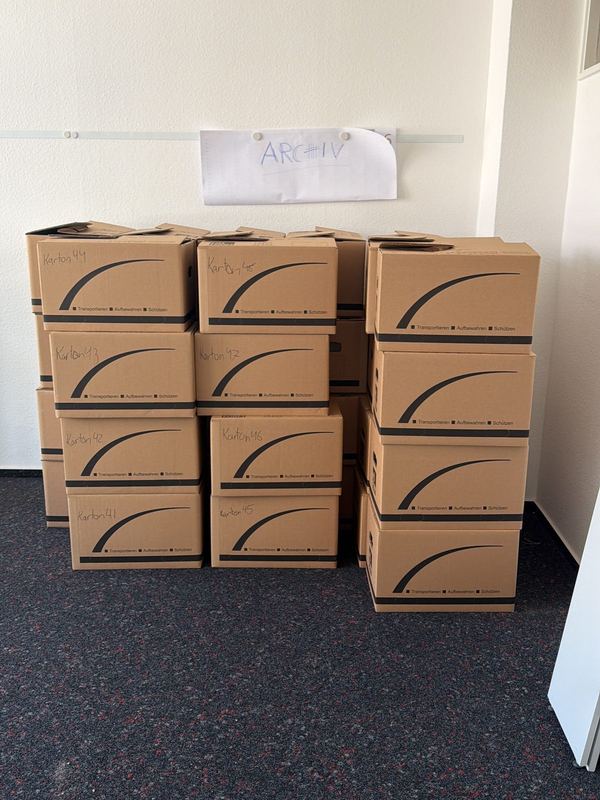Kartons nach Räumen geordnet Gestapelte Umzugskartons mit handschriftlicher Beschriftung in einem leeren Zimmer mit dunklem Teppich, an der Wand ein Zettel mit dem Wort Archiv
