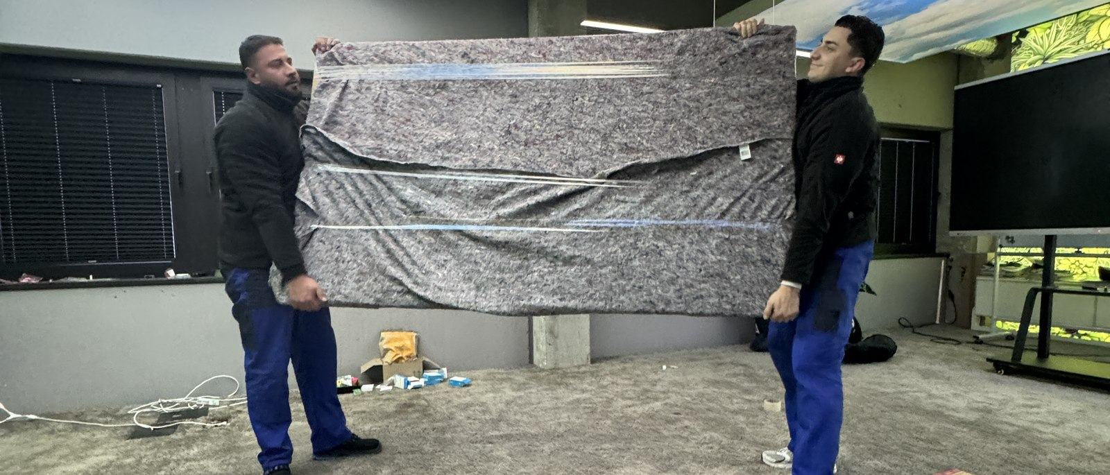 Ihr Kontakt in Meerbusch: Zwei Mitarbeiter tragen zu zweit ein großes flaches Möbelstück, das in eine graue Umzugsdecke gewickelt ist, über einen Hof an einer Hauswand entlang.