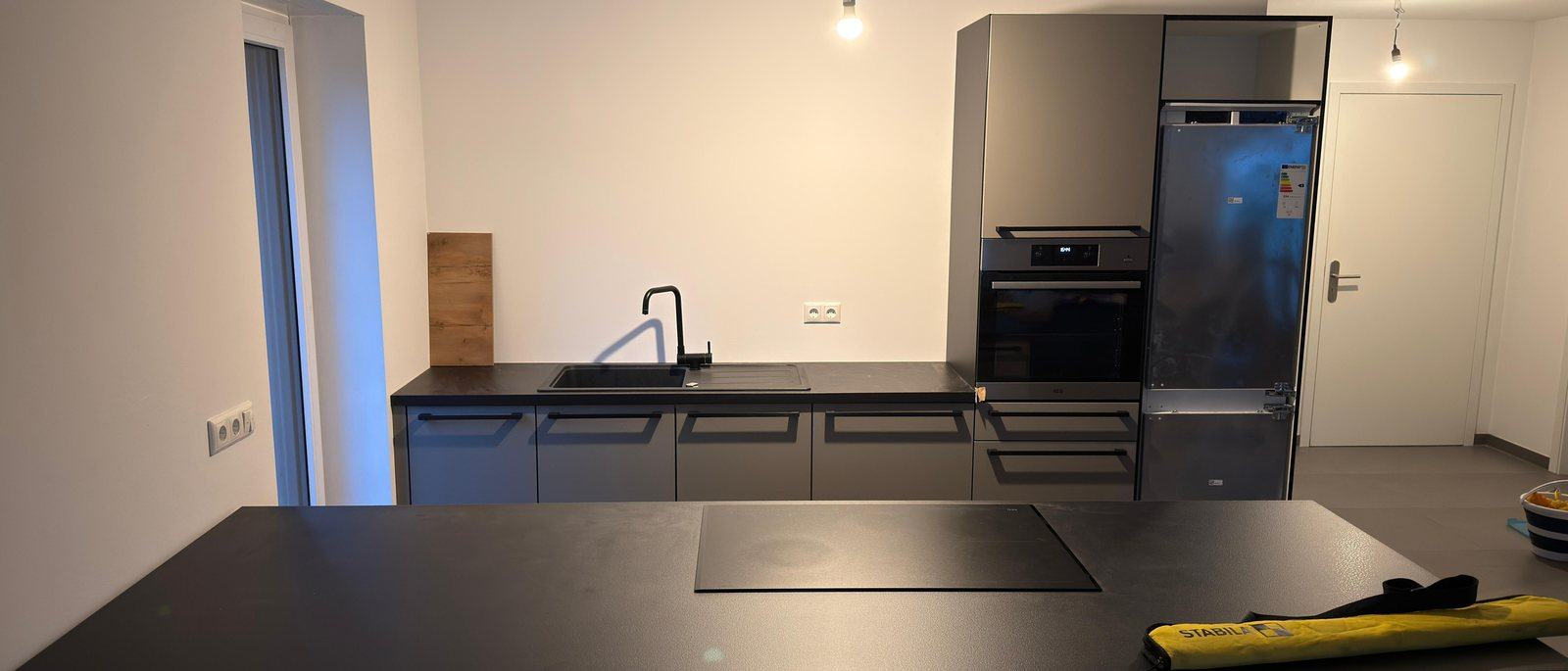 Küchenmontage in Meerbusch: Eine fertig montierte moderne Küche mit dunklen Fronten, Kochinsel mit Induktionsfeld, Spüle, Backofen und Kühlschrank in einem hellen leeren Raum, auf dem Boden liegt noch eine Werkzeugtasche.