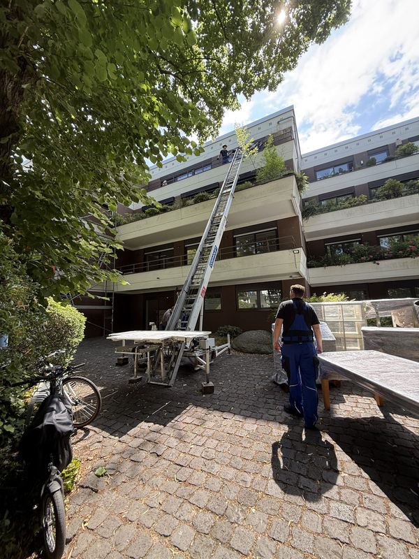 An einem mehrstöckigen Wohnhaus steht ein ausgefahrener Möbellift, unten wartet ein Mitarbeiter