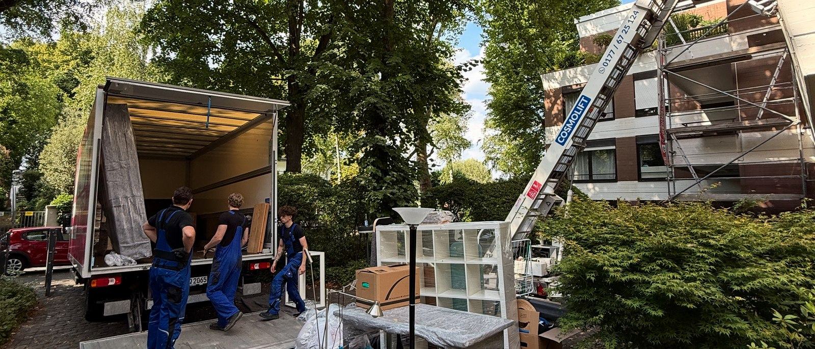 Möbellift für Umzüge in Meerbusch: Vor einem eingerüsteten Wohnhaus ist ein Möbellift bis ins obere Geschoss ausgefahren, drei Mitarbeiter stehen am geöffneten Laderaum eines Lkw neben einem Regal und Kartons