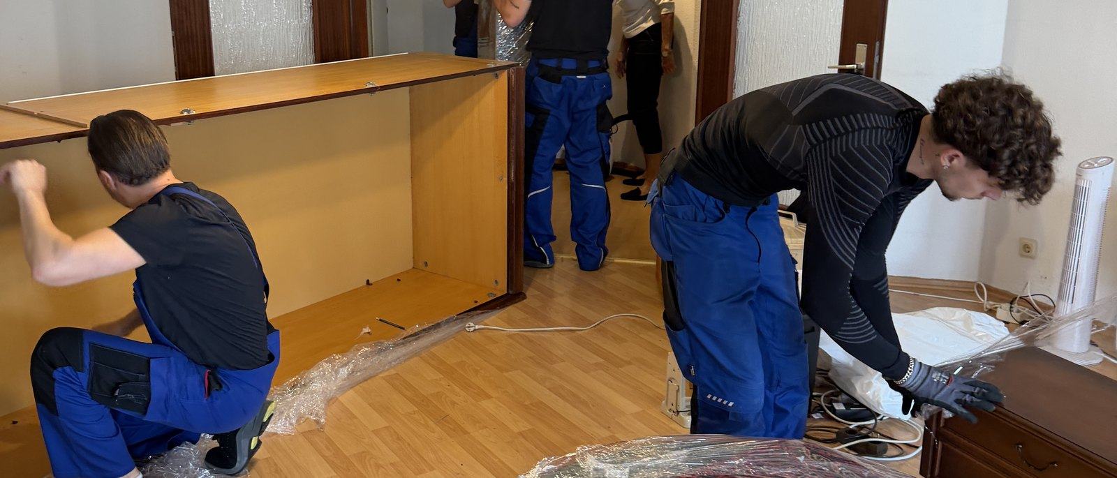 Möbelmontage in Meerbusch: Drei Mitarbeiter zerlegen in einem Wohnzimmer mit Parkett eine große Schrankwand aus Holz, die Seitenteile liegen bereits am Boden, daneben Folie und Werkzeug.