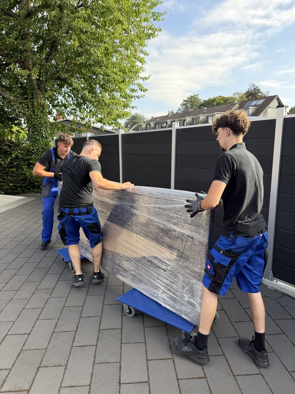 Drei Mitarbeiter rollen ein in Folie gepacktes Möbelstück auf Rollbrettern eine Einfahrt entlang