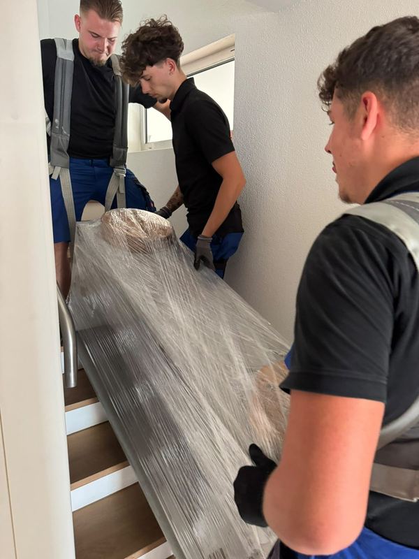 Ein in Folie gewickeltes Möbelstück wird von drei Mitarbeitern zusammen die Treppe hinuntergetragen