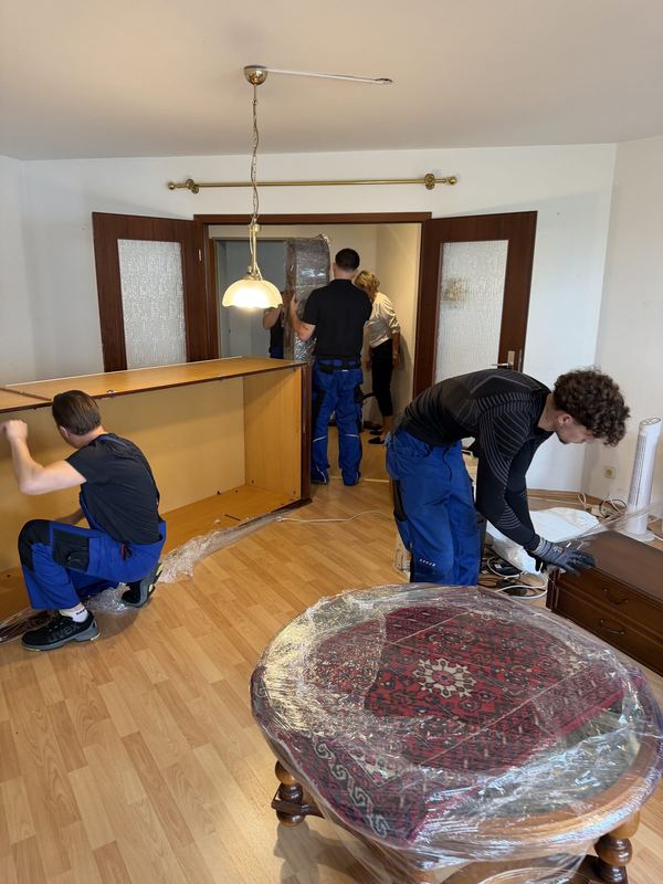 In einem Wohnzimmer stellen vier Mitarbeiter gemeinsam den Korpus einer breiten Schrankwand auf