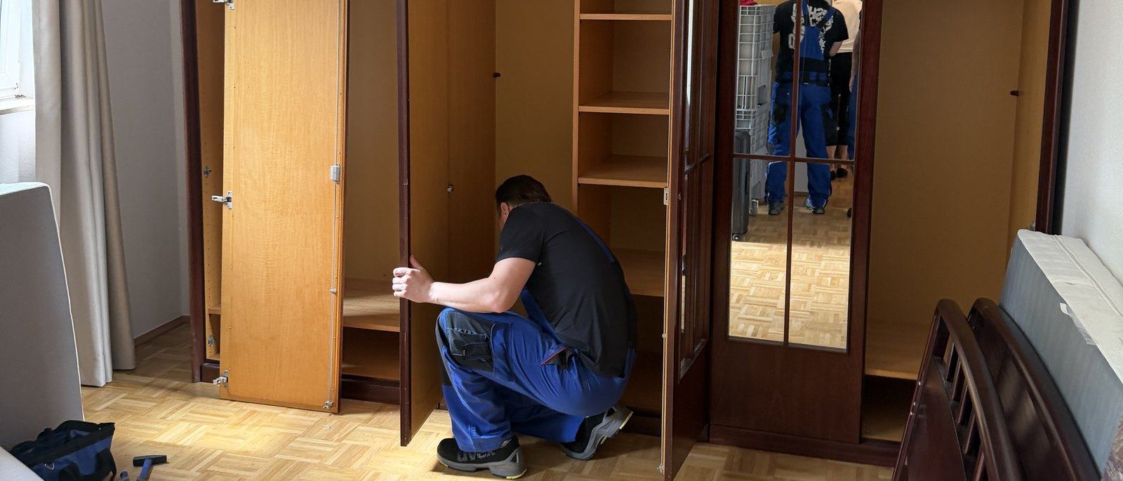 Seniorenumzug in Meerbusch: In einem Schlafzimmer mit Parkett kniet ein Mitarbeiter vor einem leer geräumten Kleiderschrank aus dunklem Holz