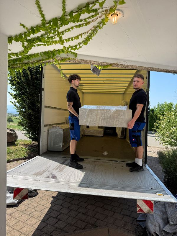 Ein foliertes Möbelstück wird von zwei Mitarbeitern über die abgesenkte Ladebordwand in den Wagen gehoben