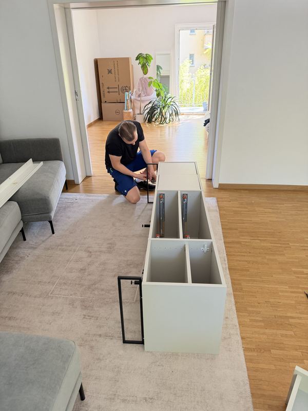 In einem hellen Wohnzimmer kniet ein Mitarbeiter vor einem weißen Sideboard und montiert es, hinter ihm stapeln sich Umzugskartons