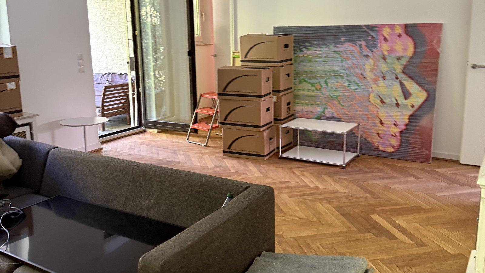 Umzugscheckliste für Meerbusch: Woche für Woche ins neue Zuhause: In einem Wohnzimmer mit Fischgrätparkett stehen gestapelte Umzugskartons neben einem Sofa und einem Beistelltisch, an der Wand lehnt ein großes buntes Bild, dahinter eine Terrassentür.
