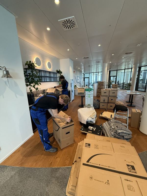 In einem Büro klebt ein Mitarbeiter einen Umzugskarton zu, dahinter stehen weitere Kartons