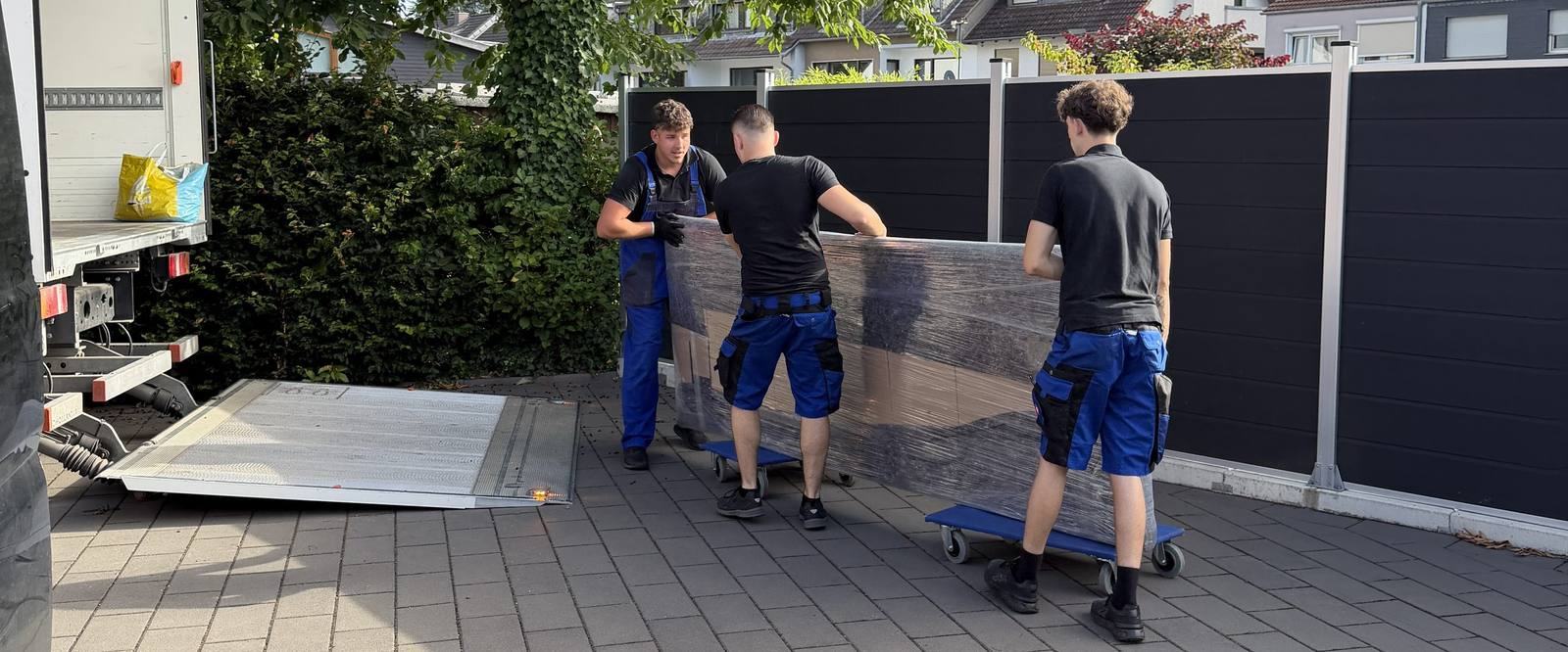 Umzugsunternehmen Meerbusch: Drei Kollegen rollen ein foliertes Möbelstück auf Rollbrettern über einen Hof zum Umzugswagen, dessen Ladebordwand abgesenkt ist