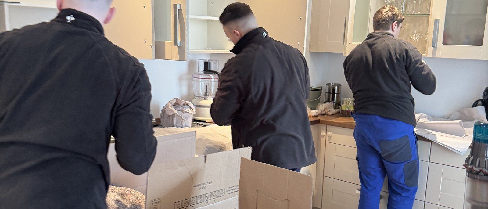 Kartons, Kleiderboxen und Schutzmaterial für Meerbusch: Drei Mitarbeiter packen in einer Küche mit hellen Fronten Geschirr und Kleingeräte in Umzugskartons auf der Arbeitsplatte, in der Mitte ein offener Karton mit Packpapier.