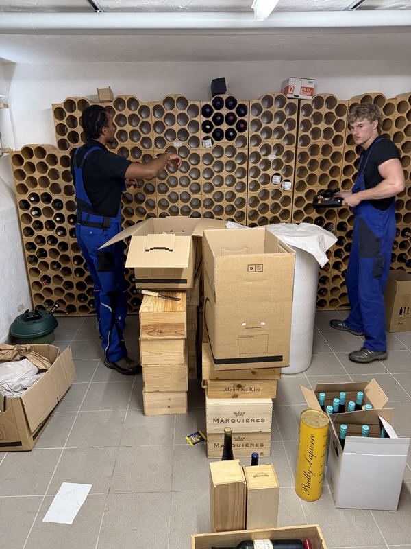 Zwei Mitarbeiter holen Wein aus einem Kellerregal aus Tonröhren und verteilen die Flaschen auf Kartons und Holzkisten
