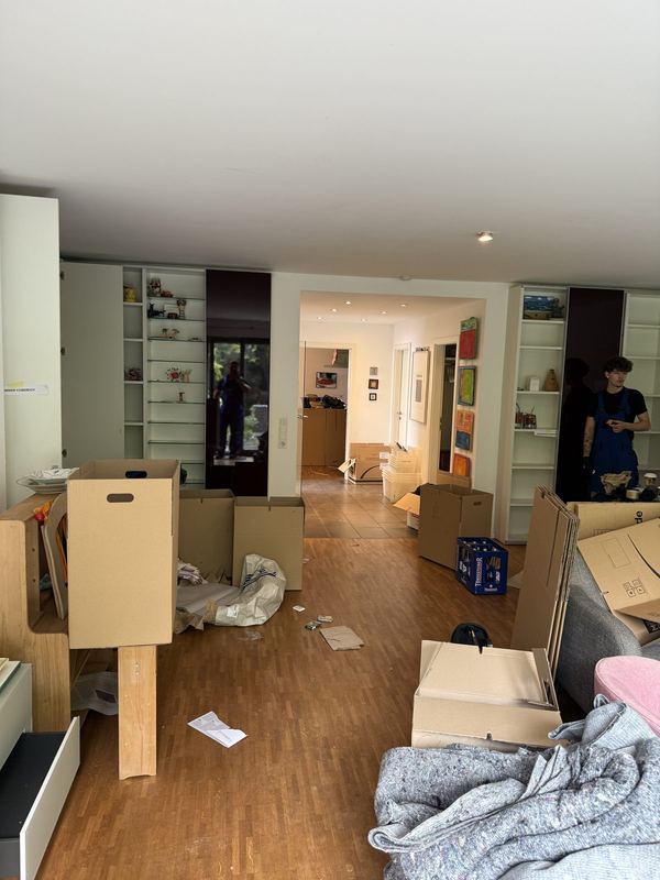 Ein Wohnzimmer mit schon geleerten Einbauregalen und aufgestellten Kartons, im Hintergrund zwei Mitarbeiter